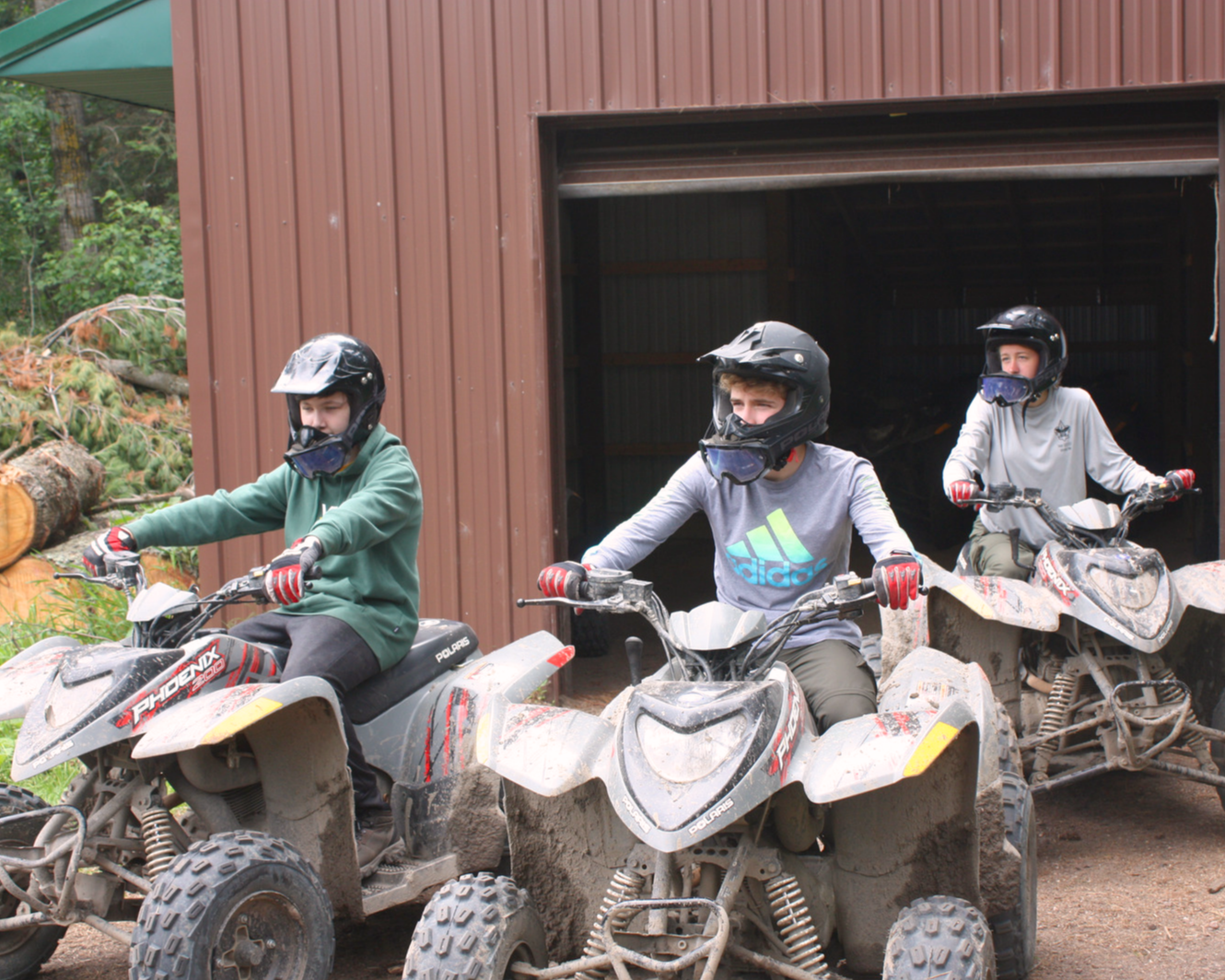 A group of Scouts wearing helmets and other protective gear are driving ATVs out of the garage they are stored in to the field
