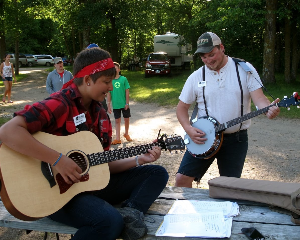 Two staff members are playing instruments while sitting on a picnic table next to the road.